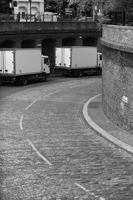 A black and white photograph showing a narrow, curved cobblestone street with three white box trucks parked in sequence along the pavement near an underpass with rounded arches. The trucks are positio