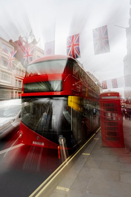 A red double-decker bus labeled 'Hammersmith' is stopped along a city street, with buildings featuring traditional architecture and a tall church spire visible in the background. The bus is part of lo