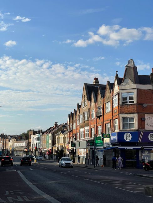 A busy street scene on Goldhawk Road in Hammersmith W6, showing a row of brick and rendered terraced houses with pitched roofs, some with bay windows. Street-level shops, including a green and blue st