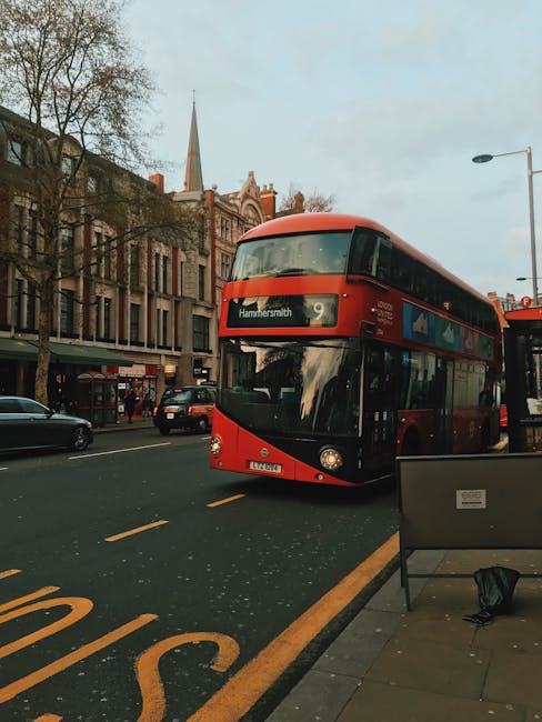 A red double-decker bus displaying the destination 'Hammersmith' is traveling along a busy street in Hammersmith, West London, near King Street flats W6. The bus is positioned in the middle of the road, with other vehicles, including cars, visible in the background. On the sidewalk beside the road, there is a black bench and a black backpack resting on the pavement. The surroundings include historic brick buildings, some with large windows, and a church steeple visible in the distance under a partly cloudy sky. The scene captures a typical urban environment during daytime, related to local transportation and city life, with the bus operated by Removal Van Hammersmith providing transport services in the area. This setting is relevant to house removals and logistical support for moving or furniture transport within the Hammersmith district.