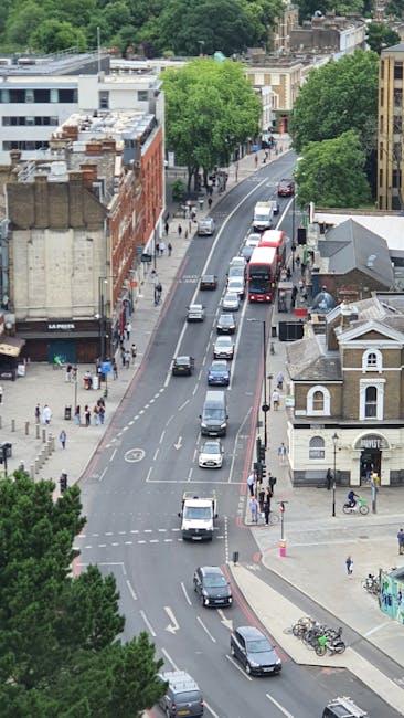 Aerial view of a busy urban street in Hammersmith, showing multiple vehicles including cars and double-decker buses traveling along a curved road. On the left side, there are several pedestrians walking on the sidewalk near shops and buildings, some waiting at a crossing. To the right, there is a small white building with an arched window and a bicycle rack with bikes parked. Tall, leafy green trees line the street, providing shade in the area. The scene captures the typical urban environment involved in home relocation and furniture transport, reflecting the logistics and movement processes typical of house removals in the Ravenscourt Park area, which Removal Van Hammersmith offers.
