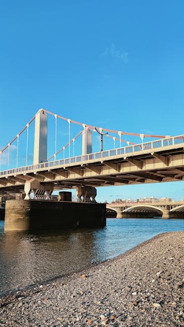 A view of Hammersmith Bridge’s suspension structure spanning the River Thames on a clear day with a bright blue sky. The bridge features tall white pylons and red suspension cables, with a pedestrian walkway and vehicle lanes visible on its deck. Below, the river reflects the bridge and the sky, with a pebble shoreline in the foreground. The scene captures the bridge’s architectural details and its location in Hammersmith, relevant for home relocation or moving logistics associated with removals services by Removal Van Hammersmith, situated near the riverside access point at Hammersmith Bridge.