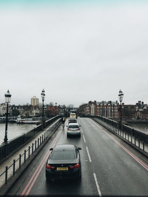 A view of a road on Hammersmith Bridge riverside under an overcast sky, with two lanes of traffic moving away from the camera. The road has metal railings on both sides, and decorative black lampposts are evenly spaced along the pavement. On the right side, a black removal van belonging to Removal Van Hammersmith is parked on the pavement, with its rear doors open, loaded with cardboard boxes, wrapped furniture, and packing materials. Some of the items are secured with straps and blankets for transportation during home relocation. The street offers scenic views of the River Thames on both sides, with historic and modern buildings visible along the riverbanks in the background. The environment appears to be part of a relocation or moving process, illustrating the logistical aspect of furniture transport and loading at this riverside location.